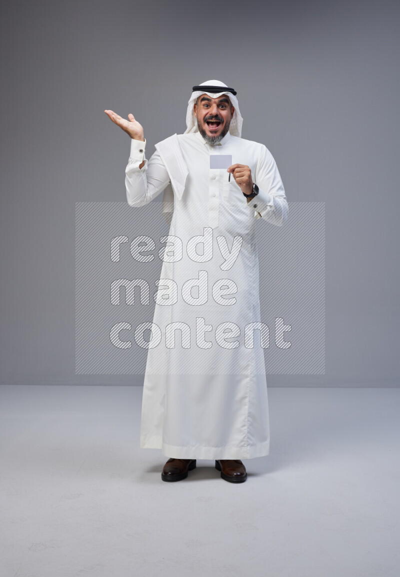 Saudi man Wearing Thob and white Shomag standing holding ATM card on Gray background