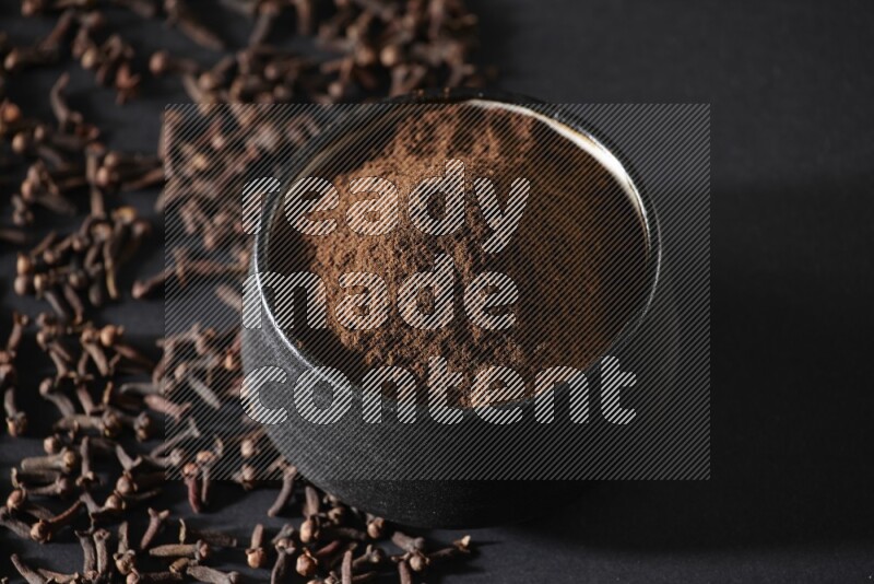 A black pottery bowl full of cloves powder and cloves whole grains on a black flooring