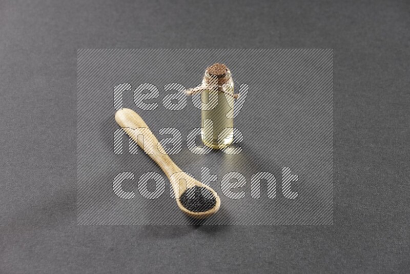 A wooden spoon full of black seeds and a glass bottle of black seeds oil on a black flooring