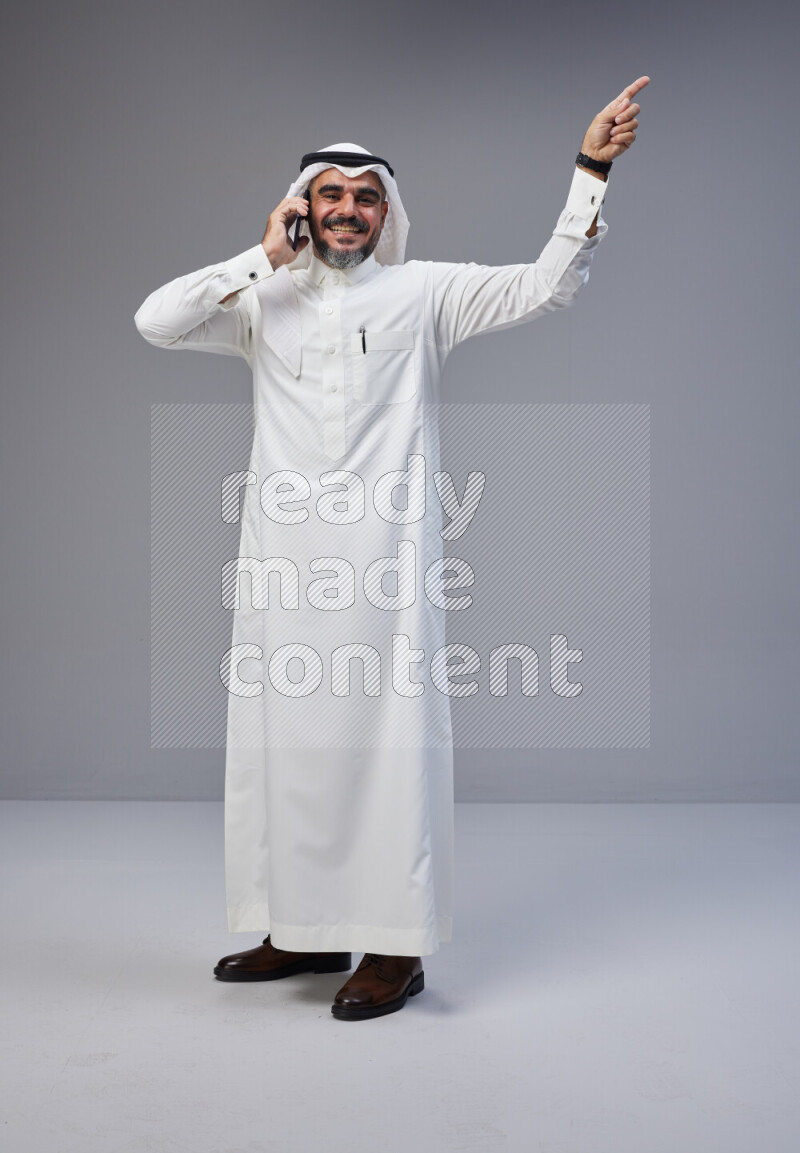 Saudi man Wearing Thob and white Shomag standing talking on phone on Gray background
