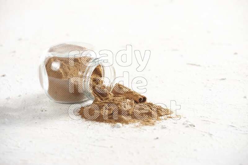 Flipped glass jar full of cinnamon powder with some pieces of cinnamon sticks on a textured white background