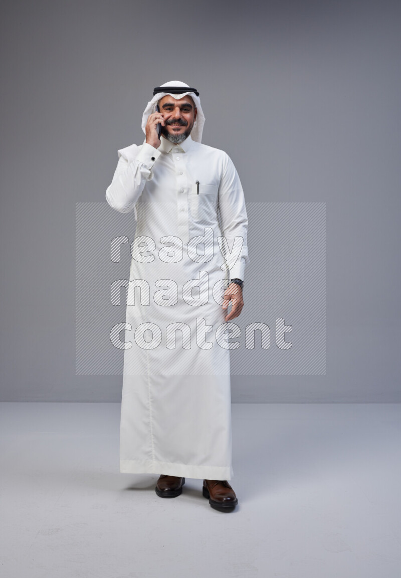 Saudi man Wearing Thob and white Shomag standing talking on phone on Gray background