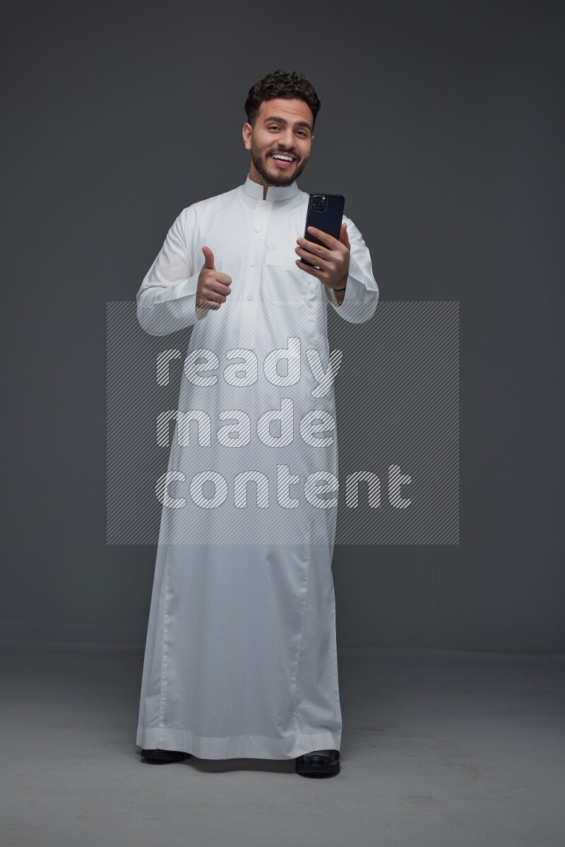 A Saudi man wearing Thobe and making a video call using his phone while standing and making different poses eye level on a gray background