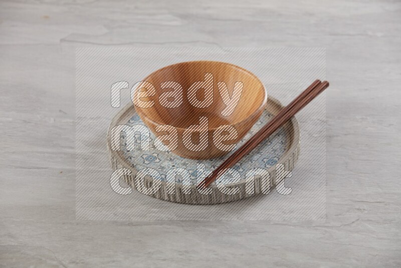 brown wood round bowl on top of multi color round ceramic plate and chopsticks, on grey textured countertop