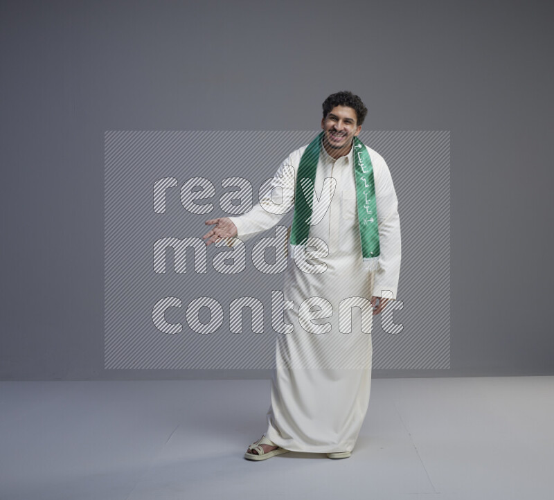 A Saudi man standing wearing thob and Saudi flag scarf on gray background