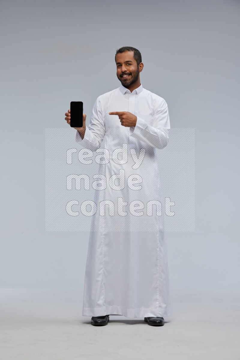 Saudi man wearing Thob standing showing phone to camera on Gray background