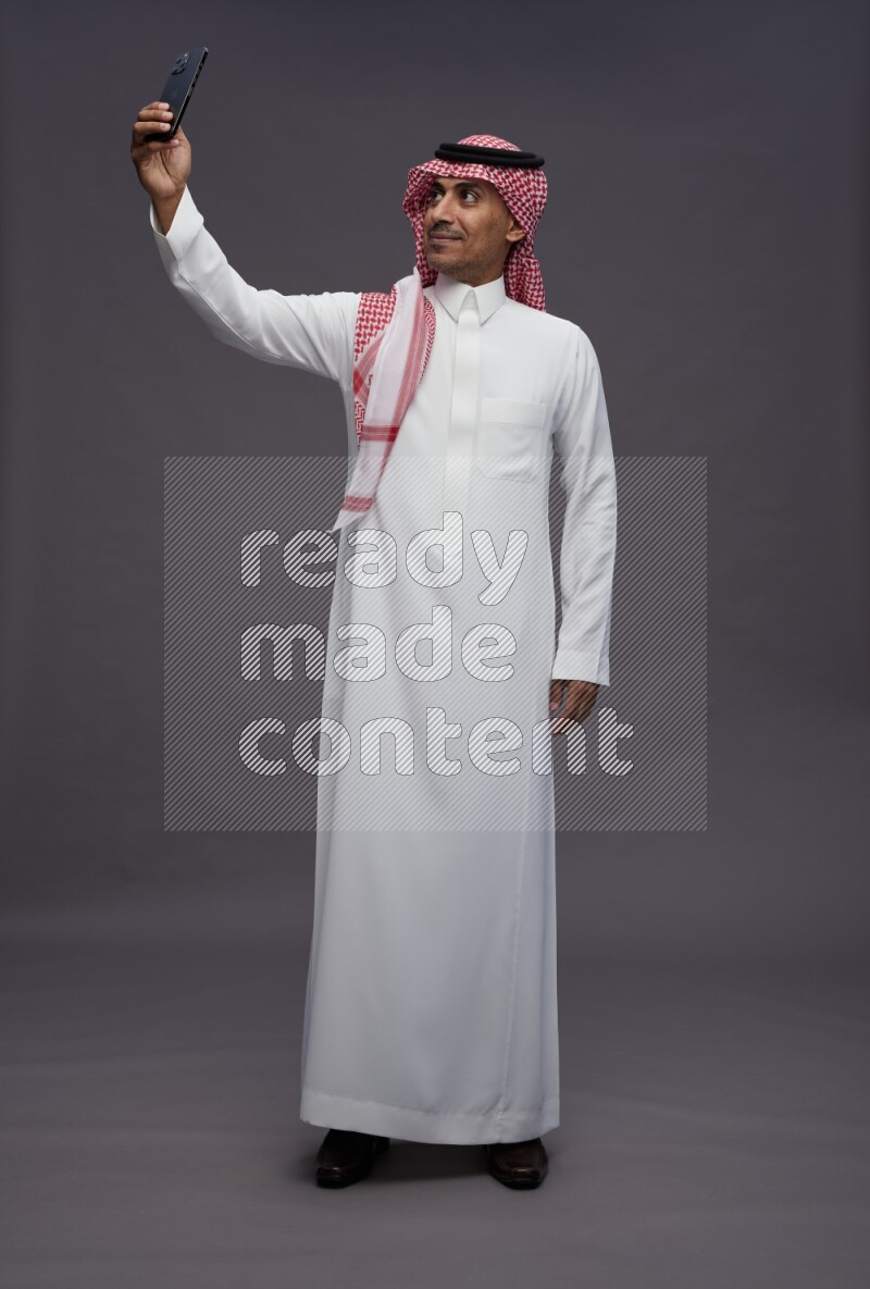 Saudi man wearing thob and shomag standing taking selfie on gray background