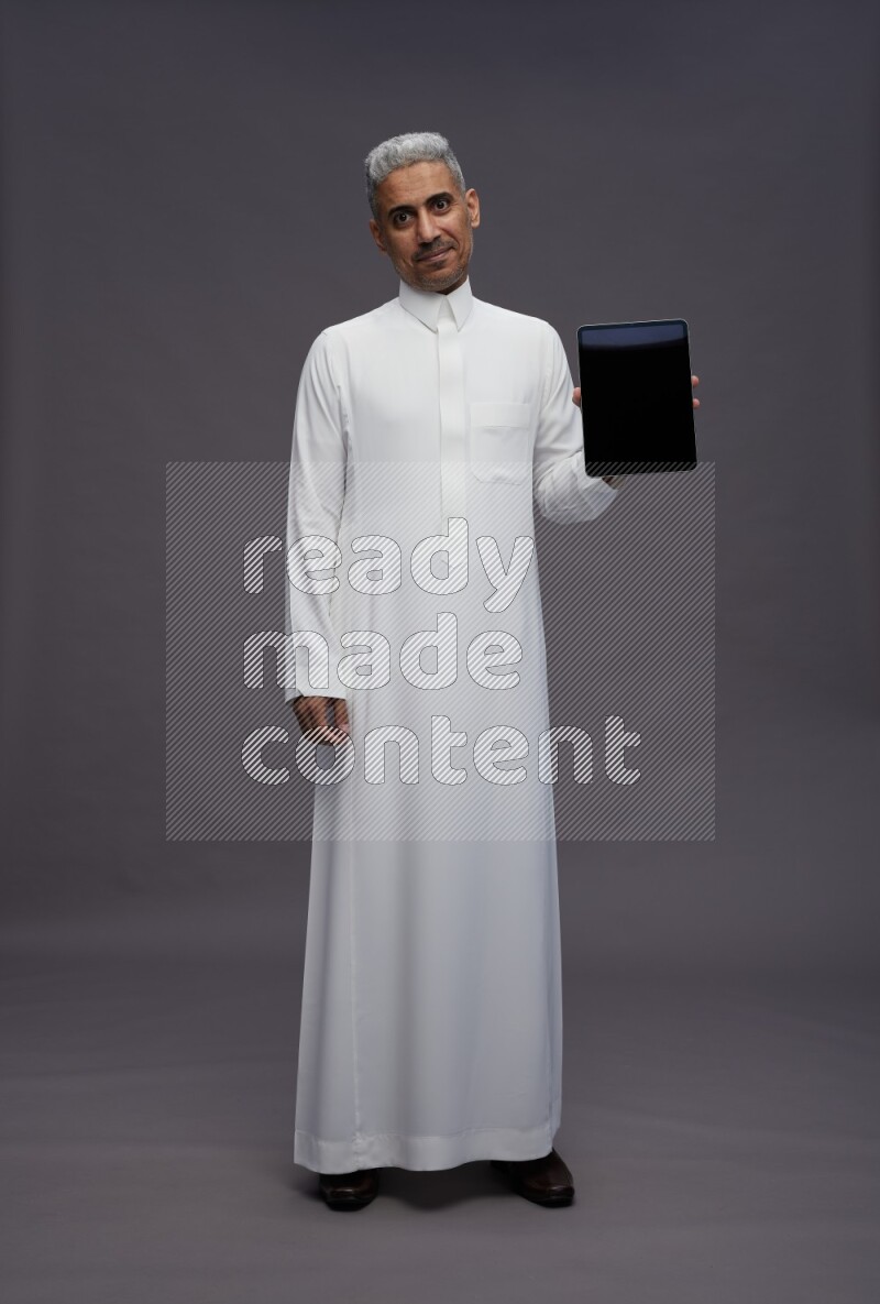 Saudi man wearing thob standing showing tablet to camera on gray background
