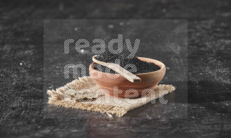 A wooden bowl full of black seeds with wooden spoon full of the seeds on it on a burlap fabric on a textured black flooring