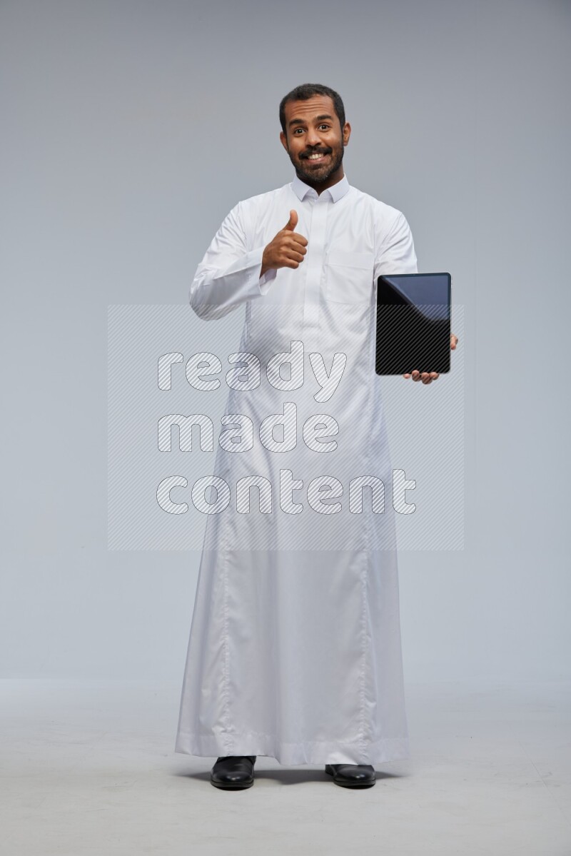 Saudi man wearing Thob standing showing tablet to camera on Gray background