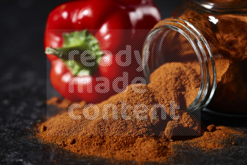 A glass jar full of ground paprika powder flipped with some spilling powder on black background