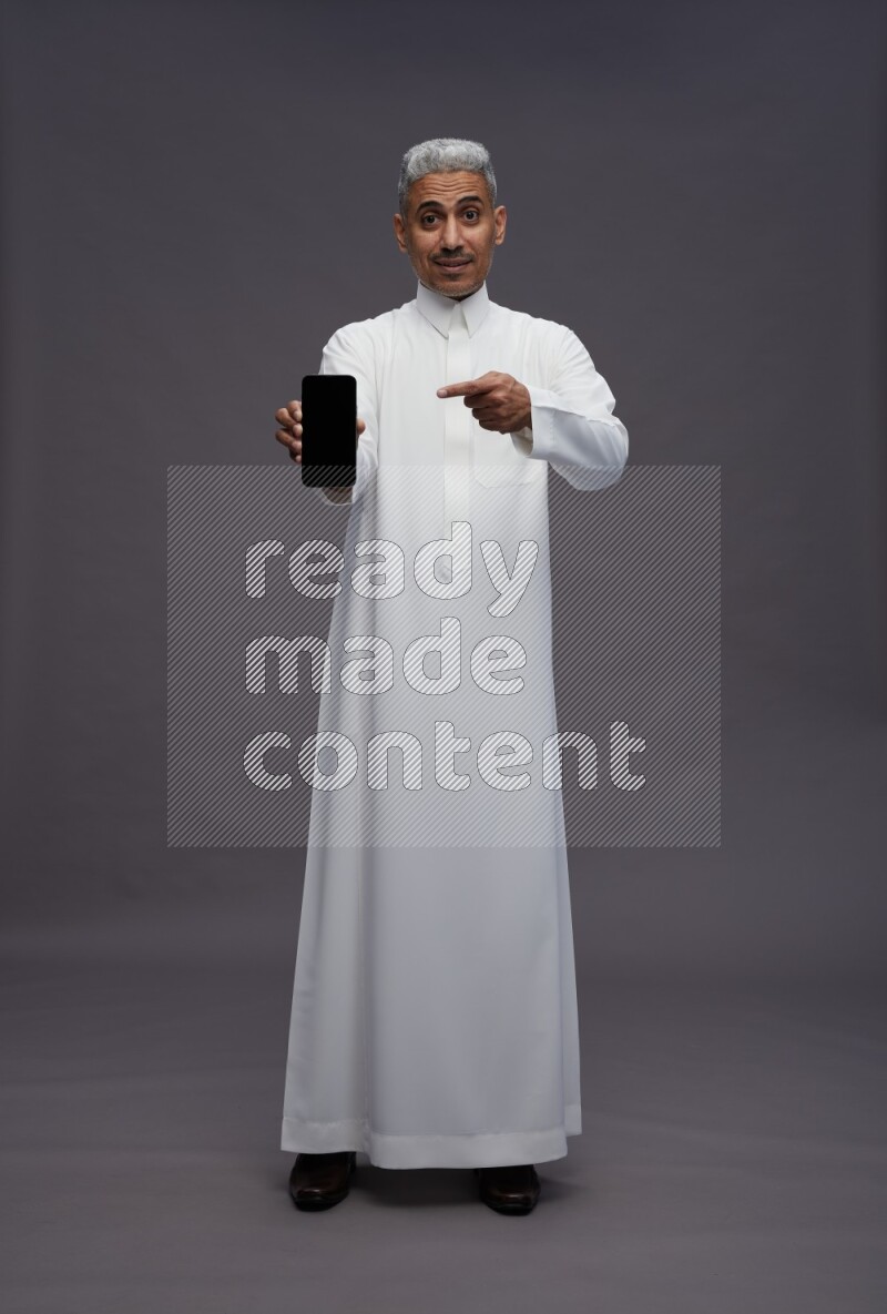 Saudi man wearing thob standing showing phone to camera on gray background