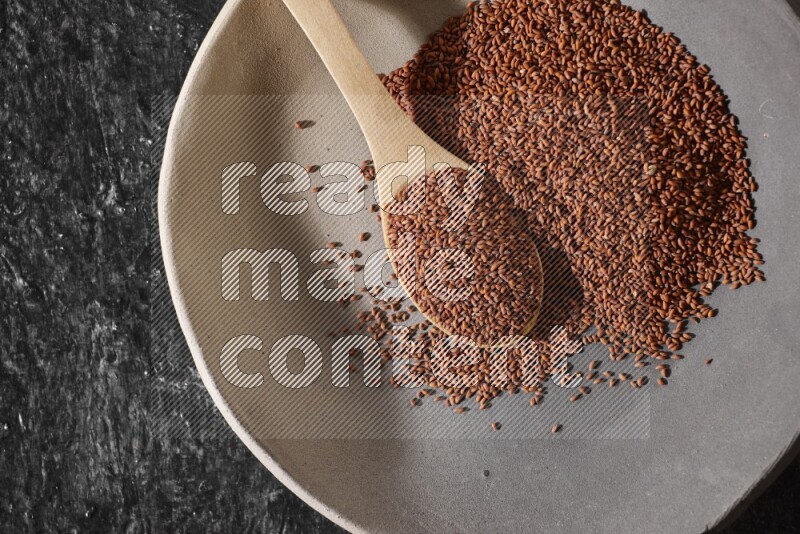 A multicolored pottery plate full of garden cress seeds and wooden spoon full of seeds on a textured black flooring