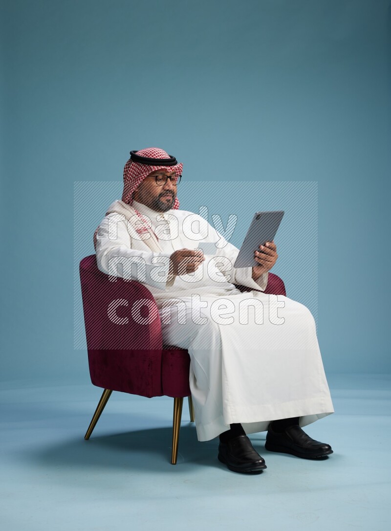Saudi Man with shimag sitting on chair holding ATM card while working on tablet on blue background