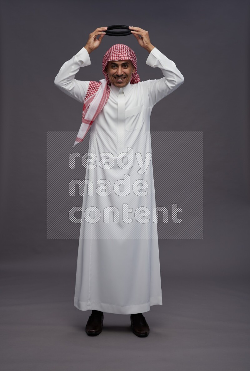 Saudi man wearing thob and shomag standing hands behind head on gray background