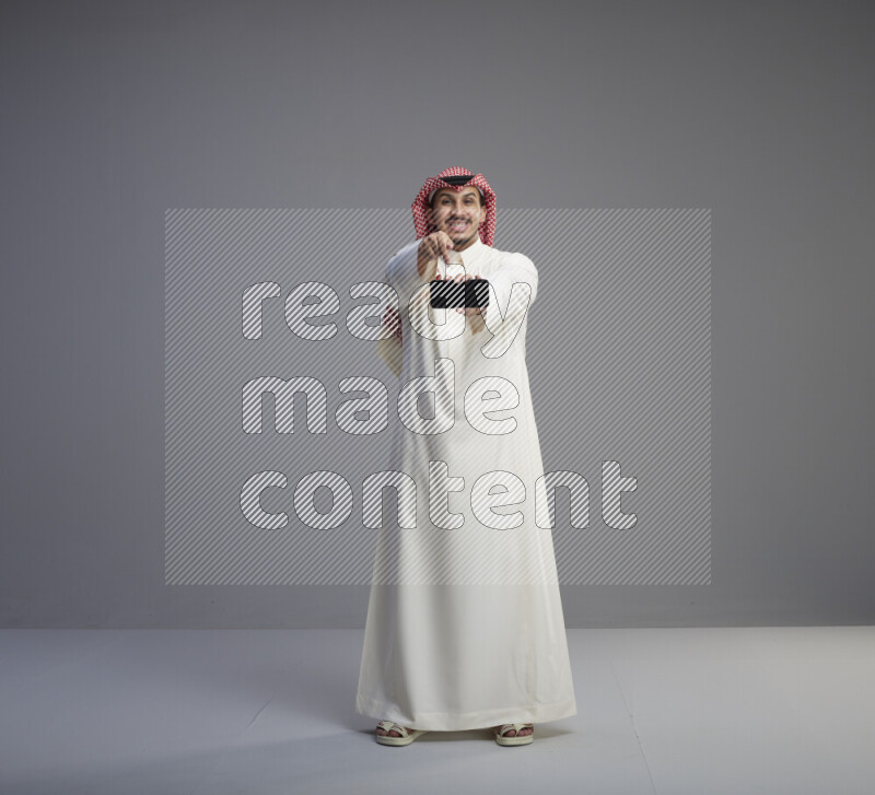 A Saudi man standing wearing thob and red shomag showing phone to camera on gray background