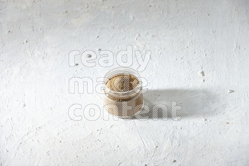 A glass jar full of cumin powder on textured white flooring