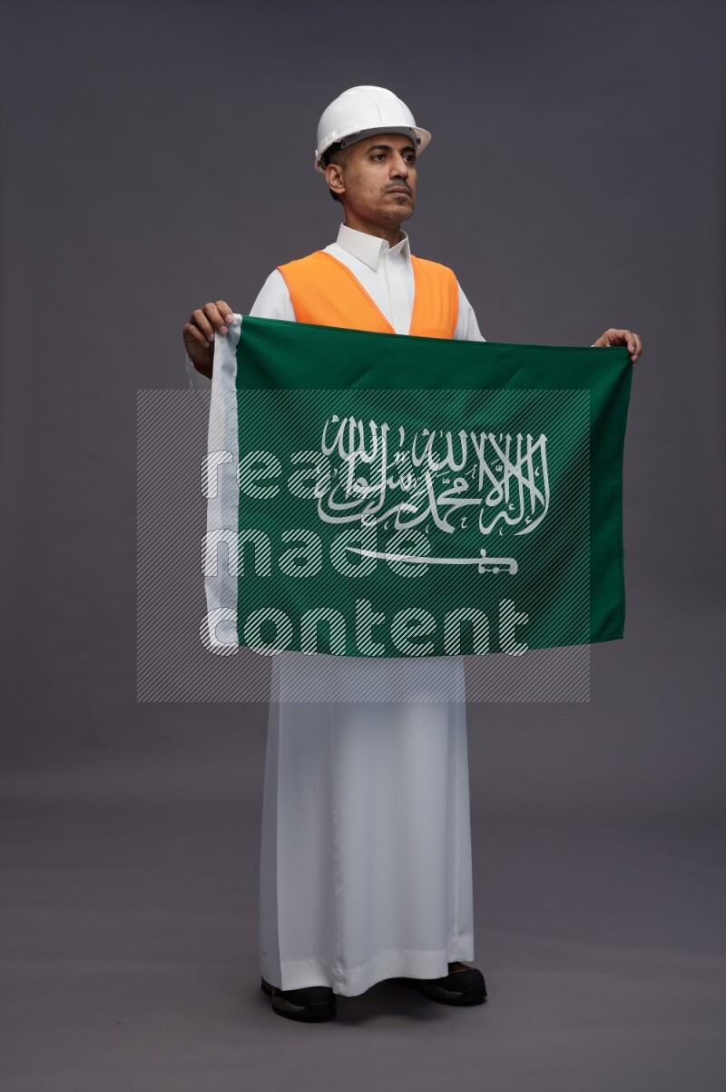 Saudi man wearing thob with engineer vest standing holding Saudi flag on gray background