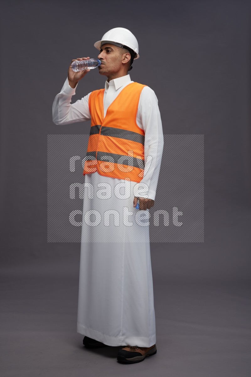 Saudi man wearing thob with engineer vest standing drinking water on gray background