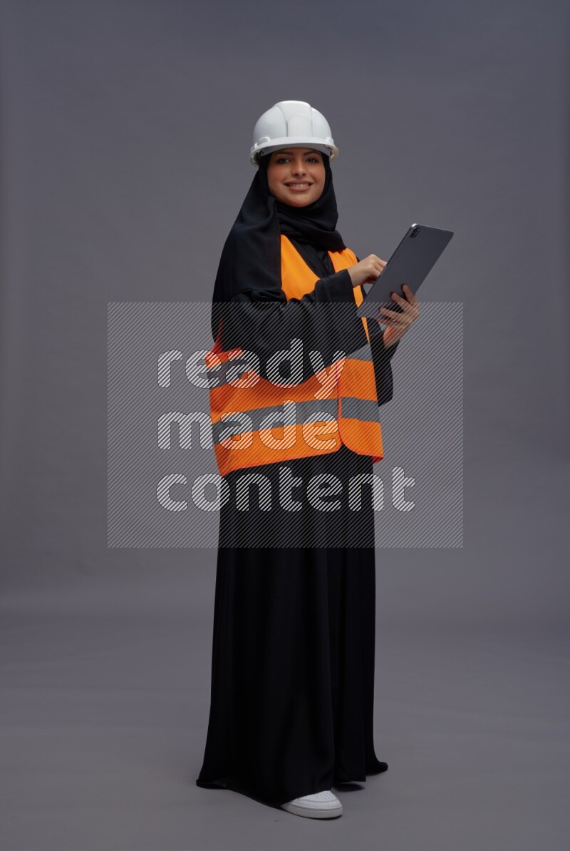 Saudi woman wearing Abaya with engineer vest standing working on tablet on gray background