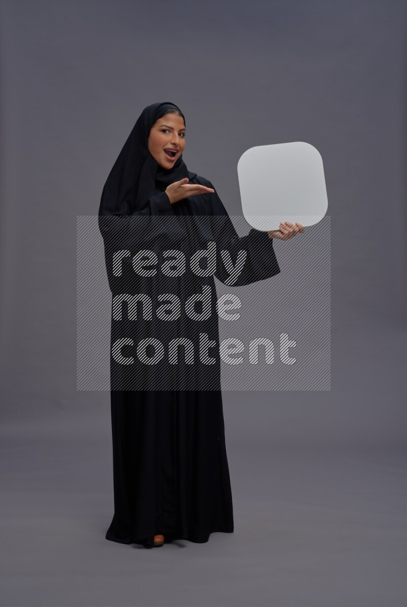 Saudi woman wearing Abaya standing holding social media sign on gray background