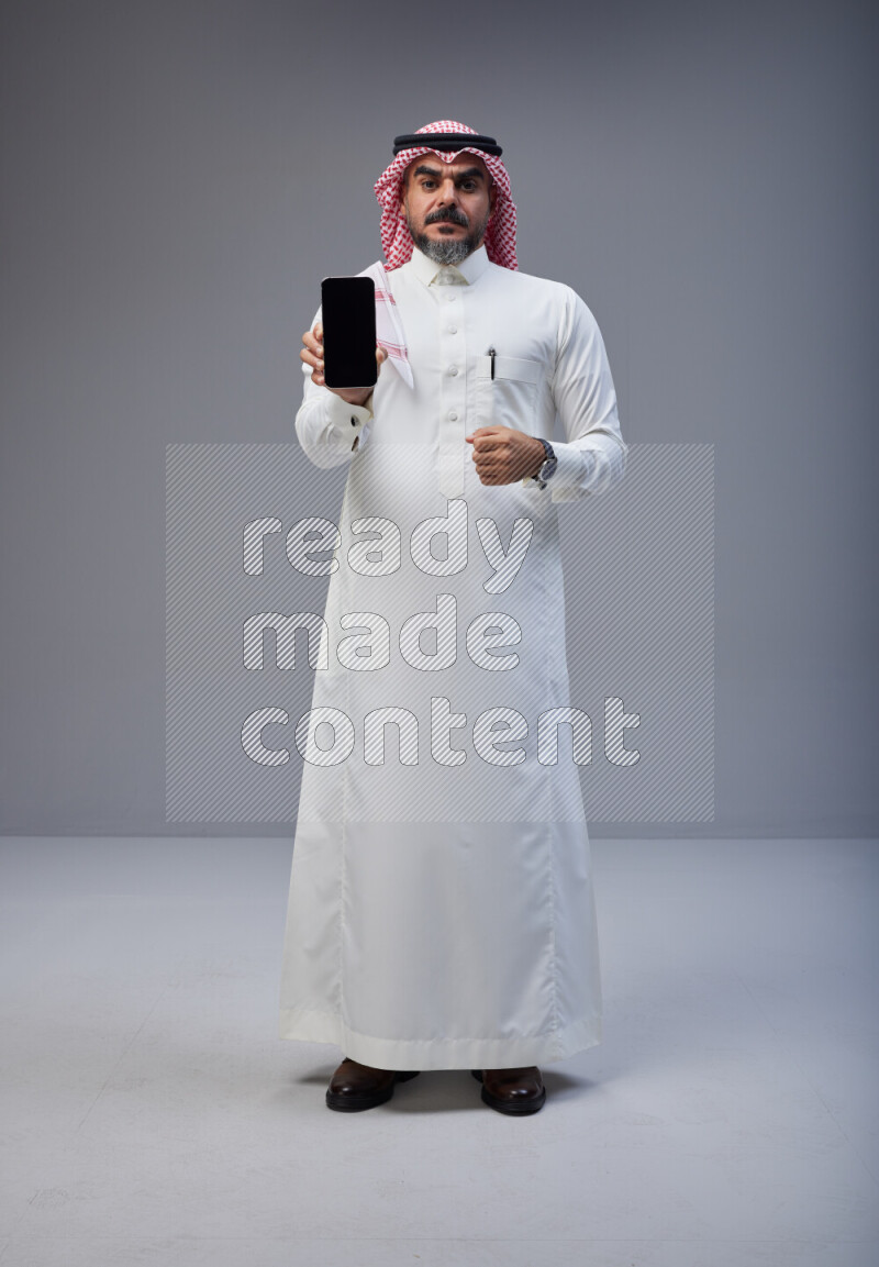 Saudi man Wearing Thob and red Shomag standing showing phone to camera on Gray background