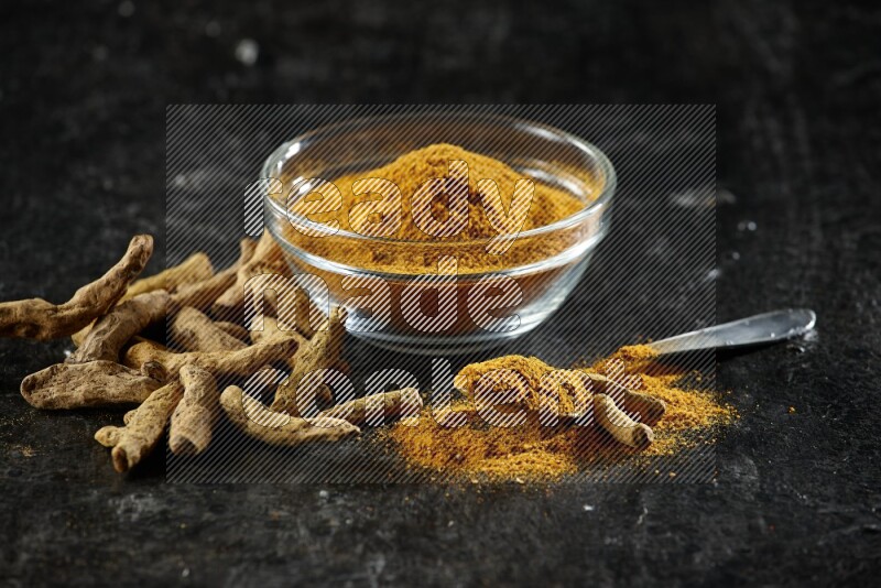 A glass bowl and a metal spoon full of turmeric powder and dried whole fingers next of them on textured black flooring