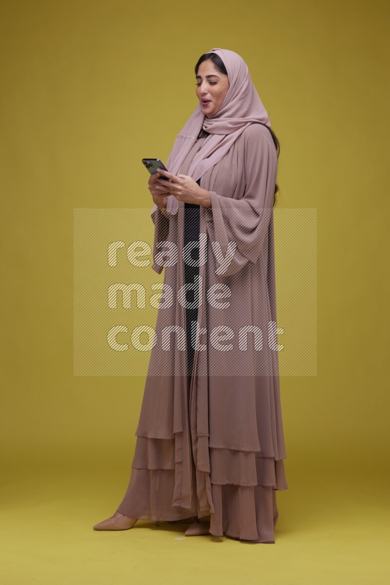 A woman Texting on her phone on a Yellow Background wearing Brown Abaya with Hijab