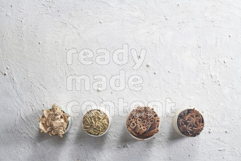 Cardamom, ginger, cinnamon sticks and star anise in 4 bowls on a textured white background