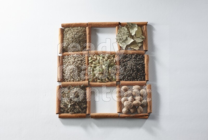 9 مربعات من أعواد القرفة ممتلئة بالهيل في المنتصف محاطة بجوزة الطيب والملح والريحان المجفف والفلفل الأبيض والكمون وبهار الفلفل الحلو وأوراق الغار والفلفل الأسود على أرضية بيضاء
