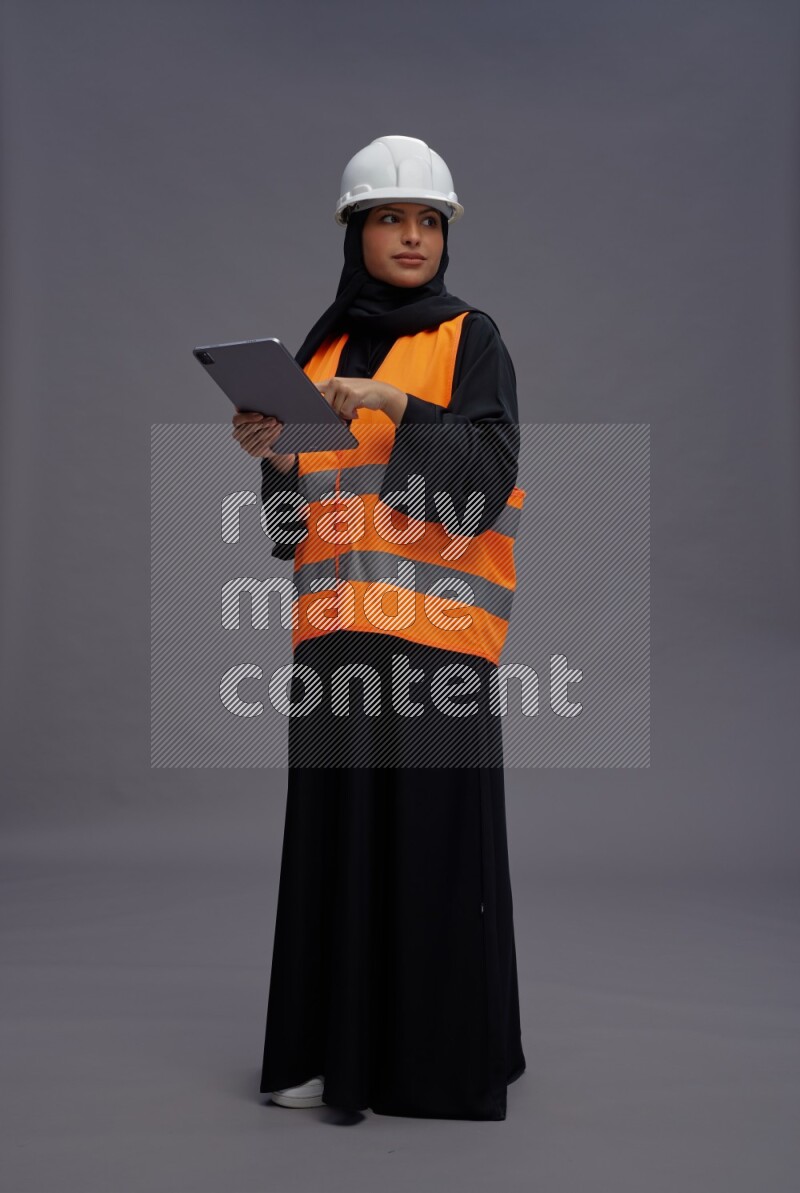 Saudi woman wearing Abaya with engineer vest standing working on tablet on gray background