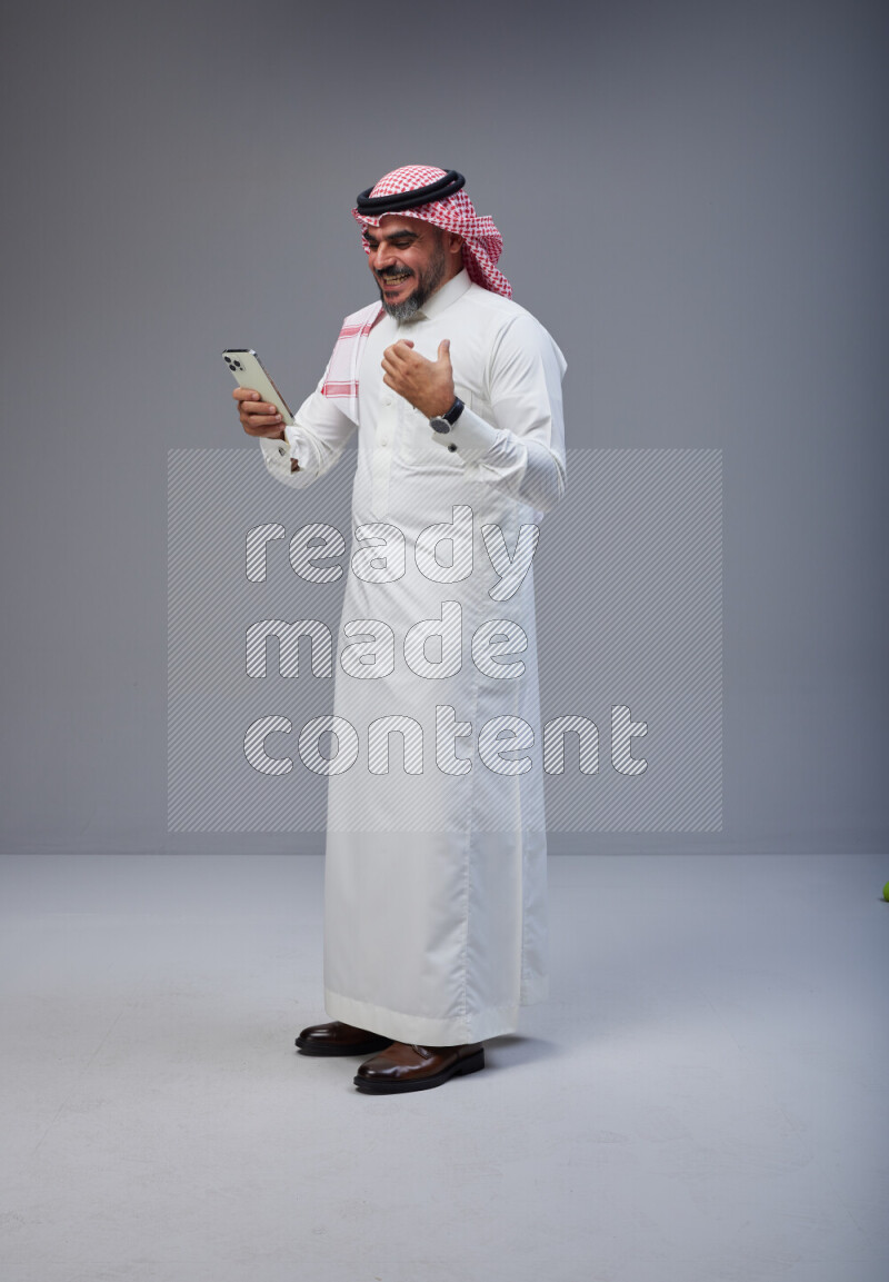 Saudi man Wearing Thob and red Shomag standing texting on phone on Gray background