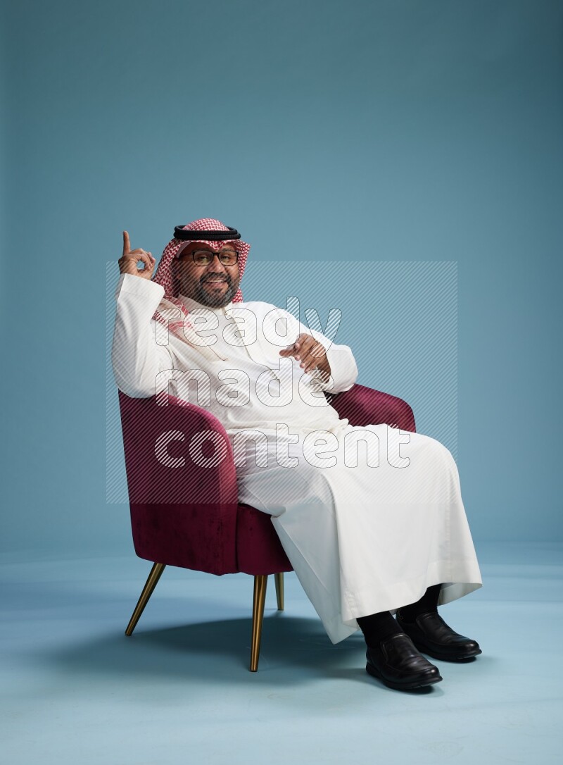 Saudi Man with shimag sitting on chair Interacting with the camera on blue background