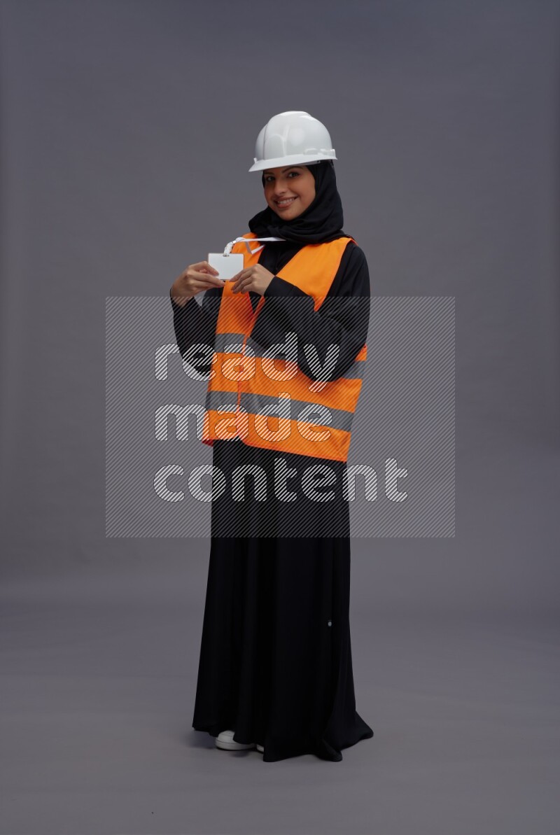 Saudi woman wearing Abaya with engineer vest with neck strap employee badge standing interacting with the camera on gray background