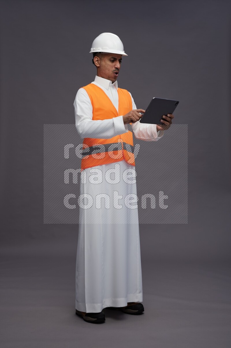 Saudi man wearing thob with engineer vest standing working on tablet on gray background