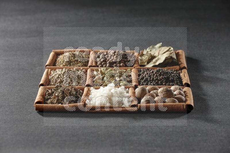 9 squares of cinnamon sticks full of cardamom in the middle surrounded by nutmeg, black pepper, bay laurel leaves, allspice, cumin, white pepper, dried basil and salt on black flooring