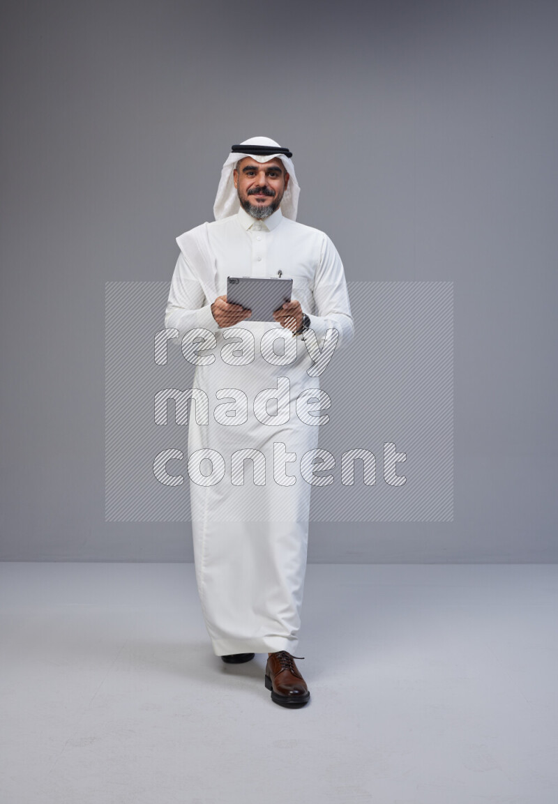Saudi man Wearing Thob and white Shomag standing working on tablet on Gray background