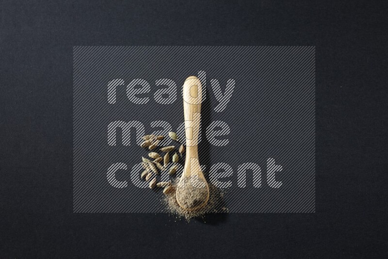 A wooden spoon full of cardamom powder and cardamom seeds beside it on black flooring