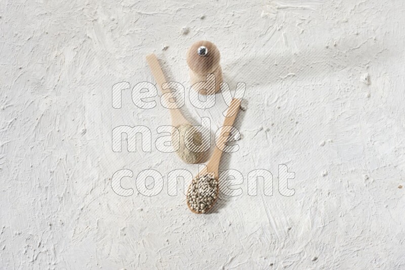 2 wooden spoons one full of white pepper powder and the other with pepper beads and a wooden pepper grinder on textured white flooring