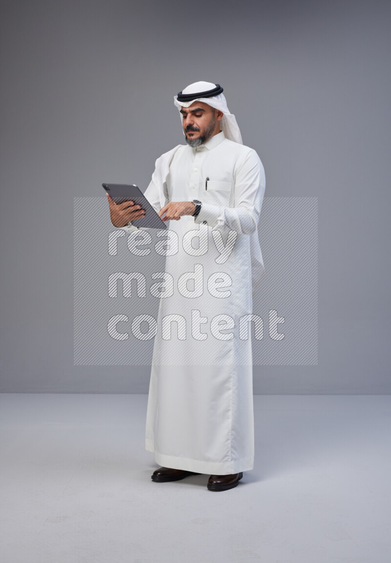 Saudi man Wearing Thob and white Shomag standing working on tablet on Gray background