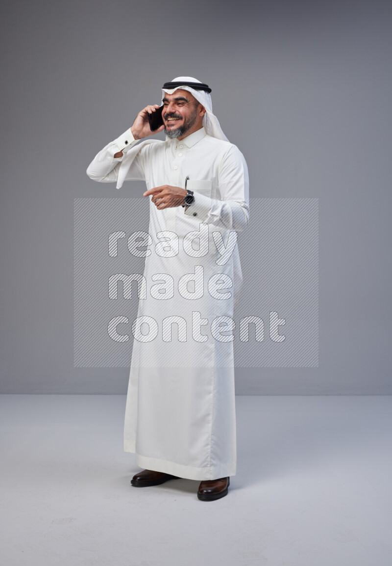 Saudi man Wearing Thob and white Shomag standing talking on phone on Gray background