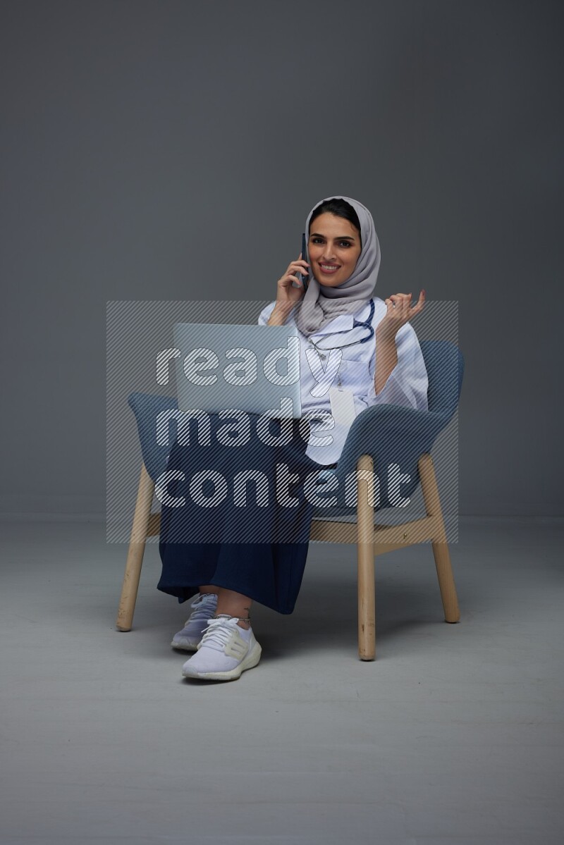 A doctor wearing a light gray head scarf sitting on blue chair on grey background
