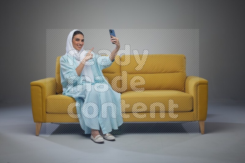 A Saudi woman wearing a light blue Abaya and white head scarf sitting on a yellow sofa and taking selfies with her phone eye level on a grey background