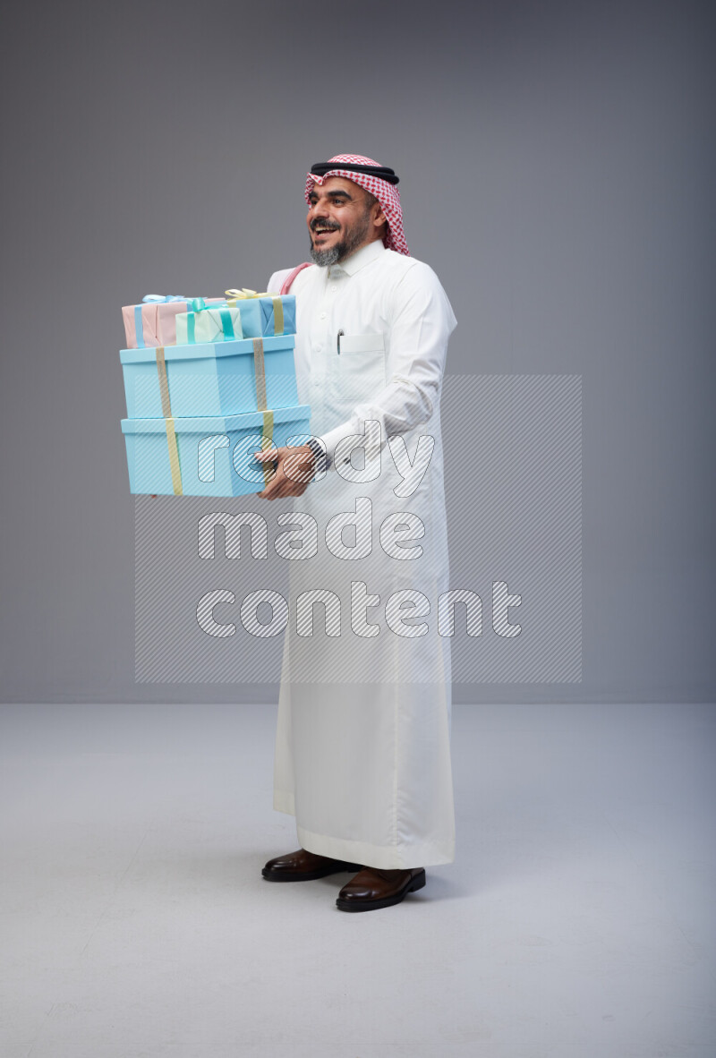 Saudi man Wearing Thob and red Shomag standing holding gift box on Gray background