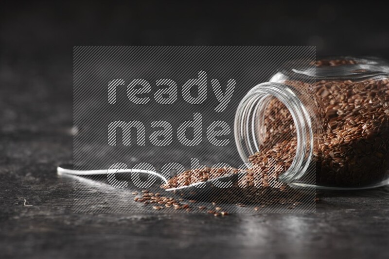 A glass spice jar full of flaxseeds flipped and seeds spread out with a metal spoon full of the seeds on a textured black flooring