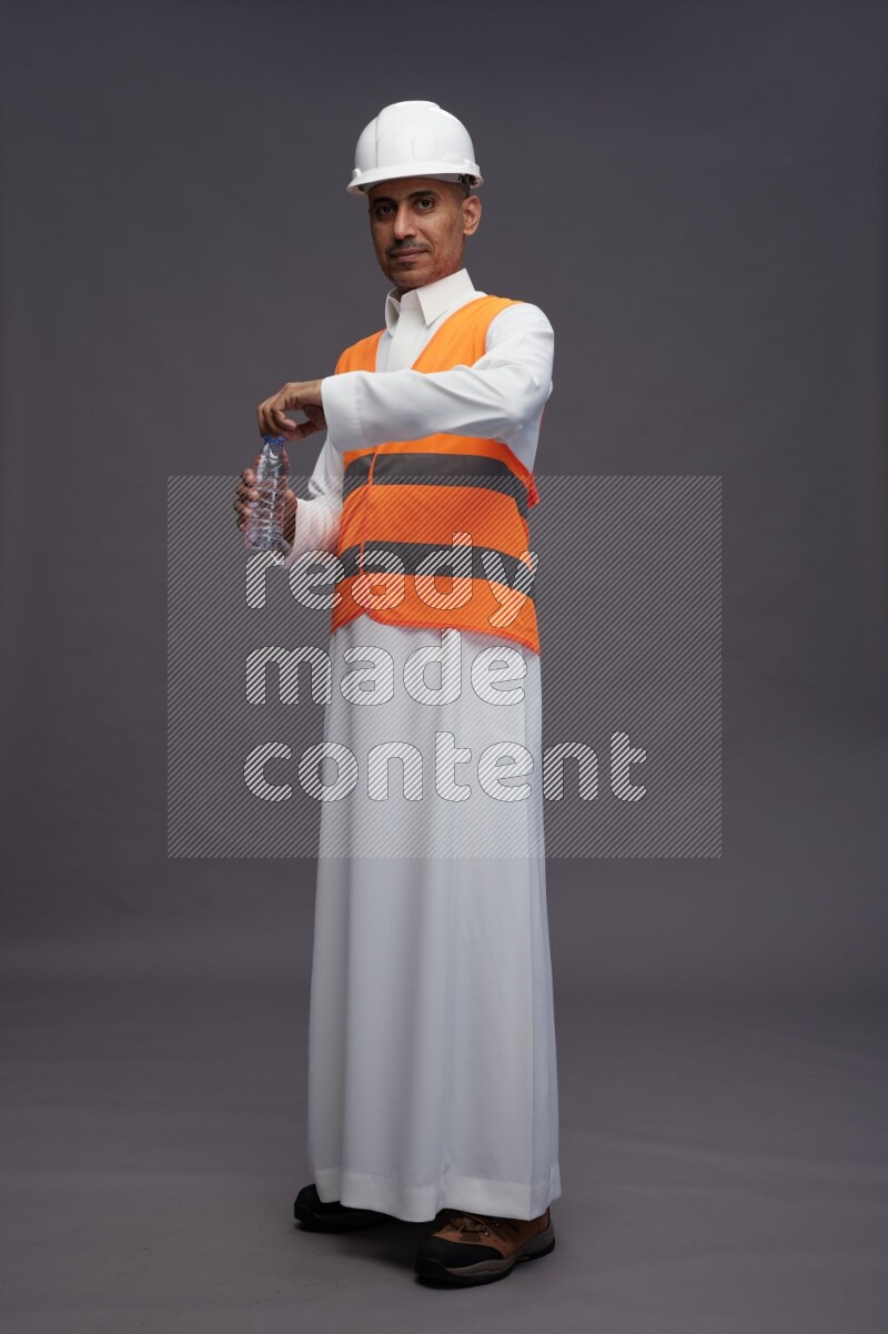 Saudi man wearing thob with engineer vest standing drinking water on gray background