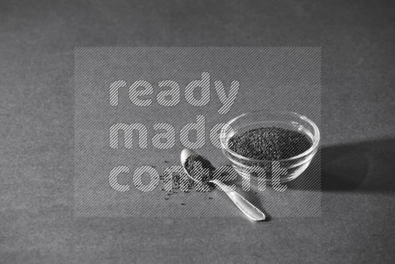 A glass bowl full of black seeds with a metal spoon full of the seeds on a black flooring