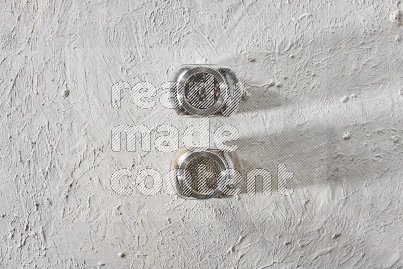 2 glass spice jars full of black pepper and black pepper powder on textured white flooring