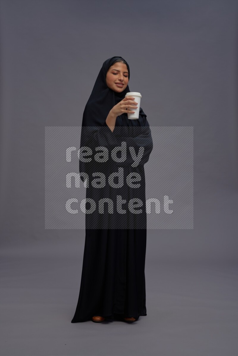 Saudi woman wearing Abaya standing holding paper cup on gray background