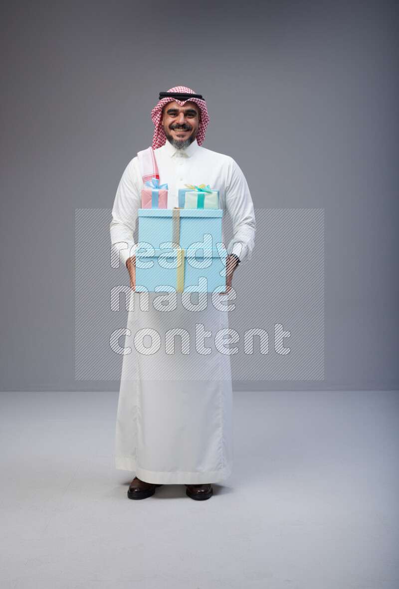 Saudi man Wearing Thob and red Shomag standing holding gift box on Gray background